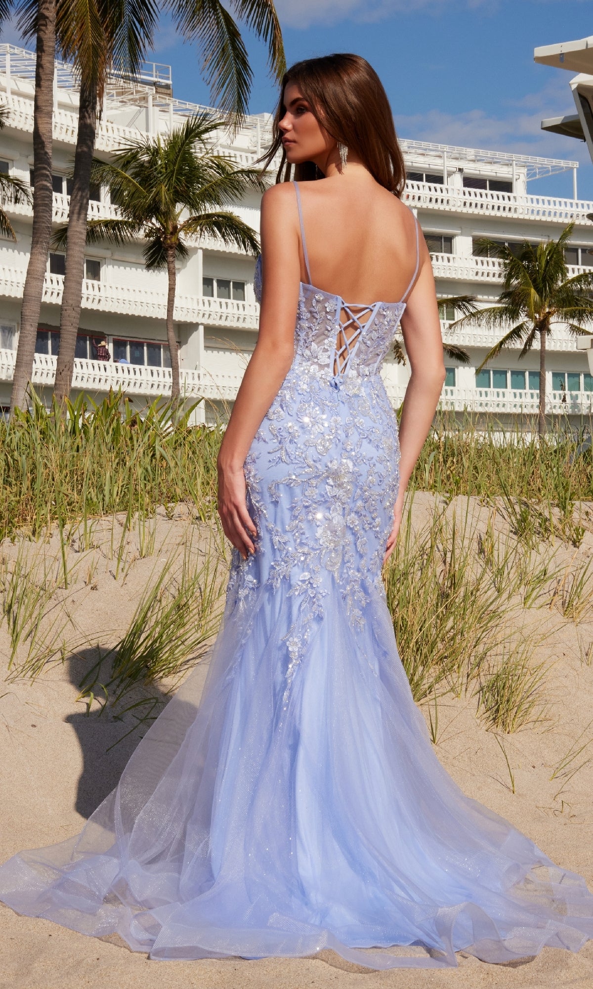 A woman stands on a sandy beach, facing away from the camera, wearing the Nox Anabel Q1541 long embroidered prom dress in light blue. Palm trees and a white building appear in the background.